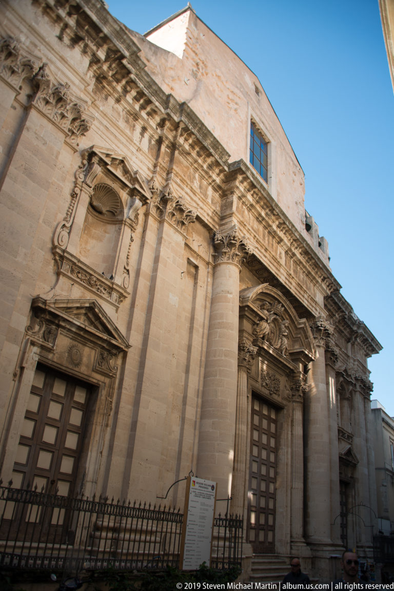 Large Baroque limestone church façade with giant pilasters, heavy cornices, and two richly carved doorways.