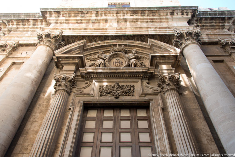 Ornate Baroque church portal with paired columns, carved floral decoration, Corinthian details, and an IHS Christogram above the doorway.