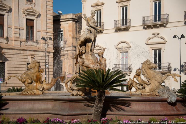 Fountain of Diana with central figures and sea creatures around the basin, partly obscured by a foreground palm.
