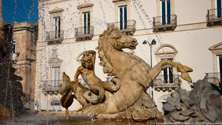 Rearing stone marine horse with attendant figure and curling forms amid fountain spray and surrounding balconies.