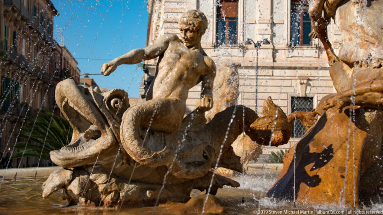 Muscular stone male figure beside an open-mouthed sea creature, crossed by multiple fountain jets.