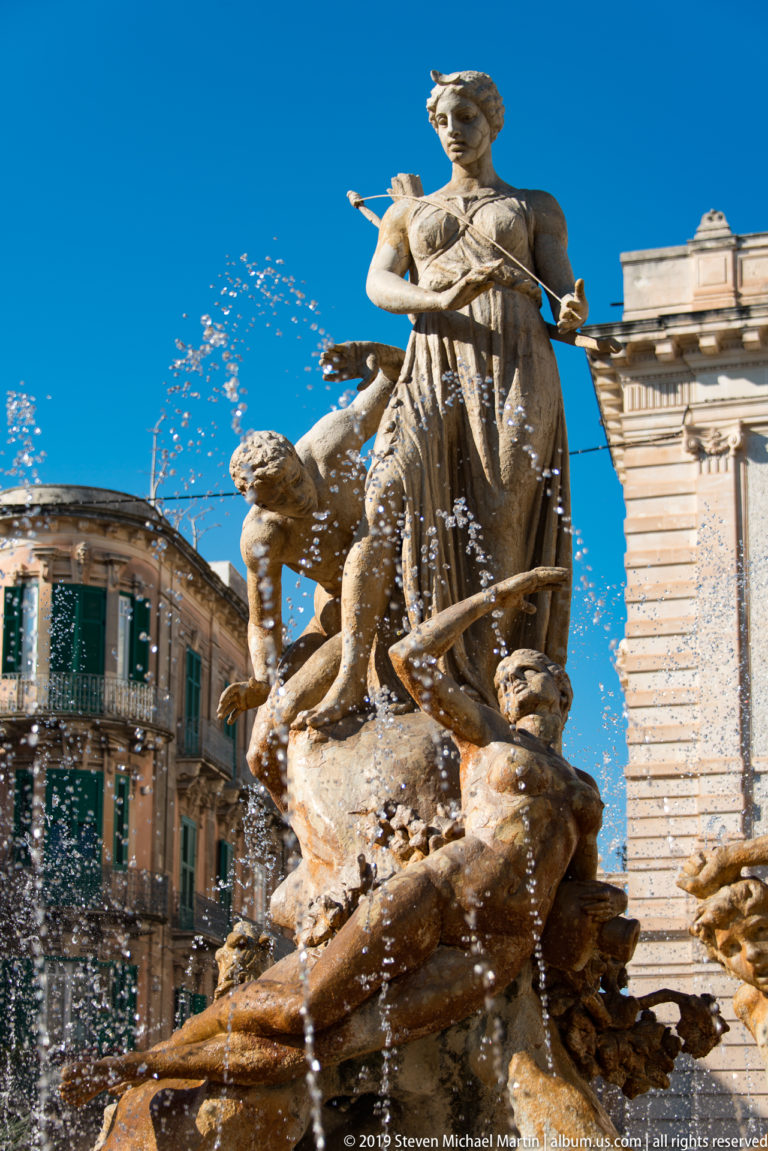 Central female statue of Diana above rocky fountain sculpture and attendant figures, with water jets crossing a blue sky.
