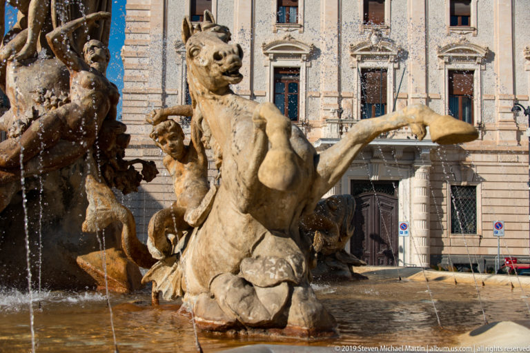 Rearing stone sea horse with curling tail surrounded by smaller figures and fountain jets in Piazza Archimede.