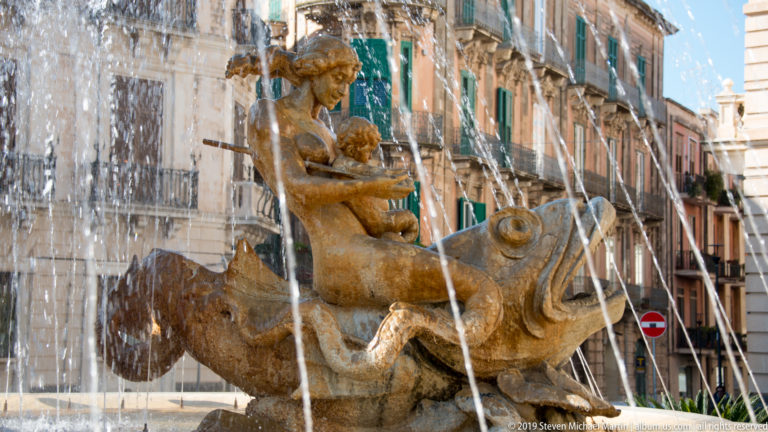 Female stone figure riding a large fish-like sea creature amid fountain jets, with historic buildings behind.