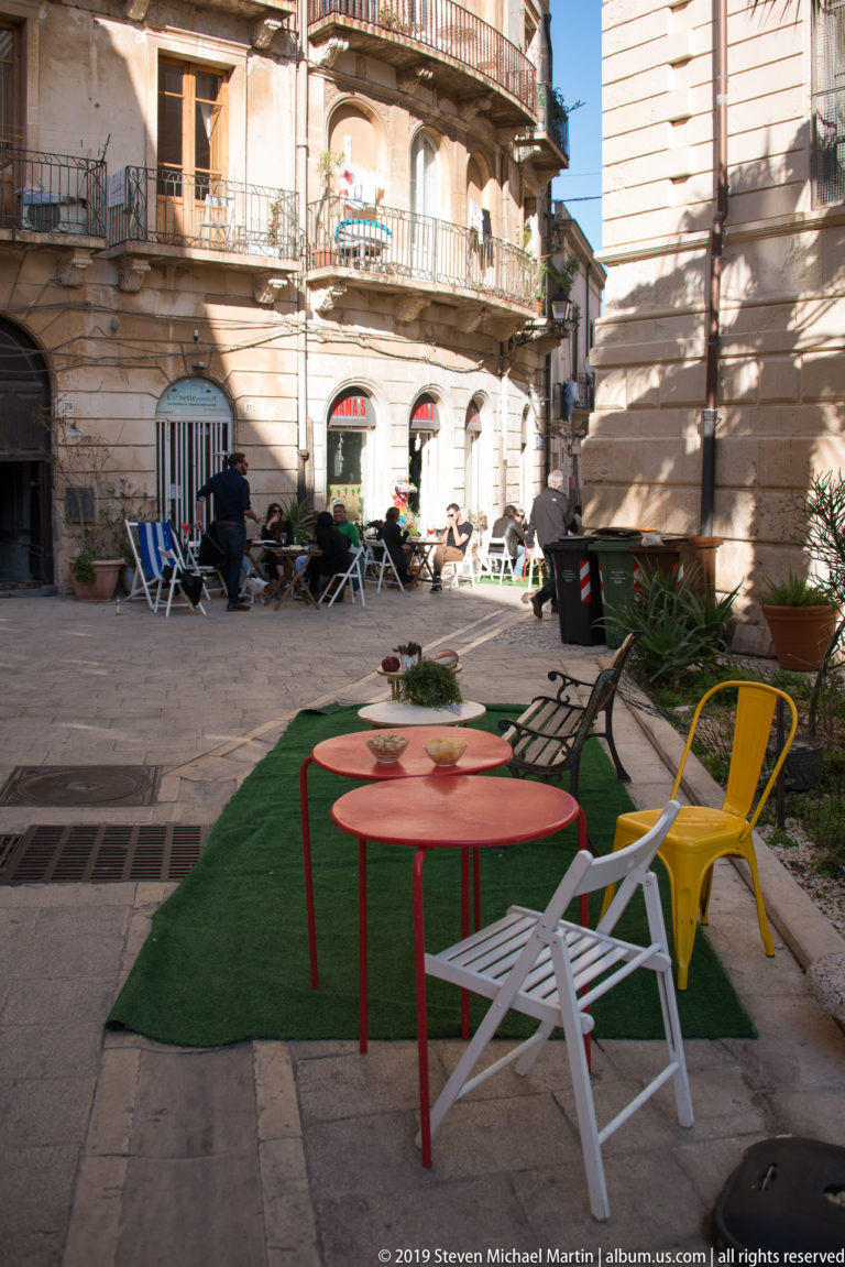 Colorful chairs and small tables arranged on paving and green turf within a historic Ortigia courtyard.