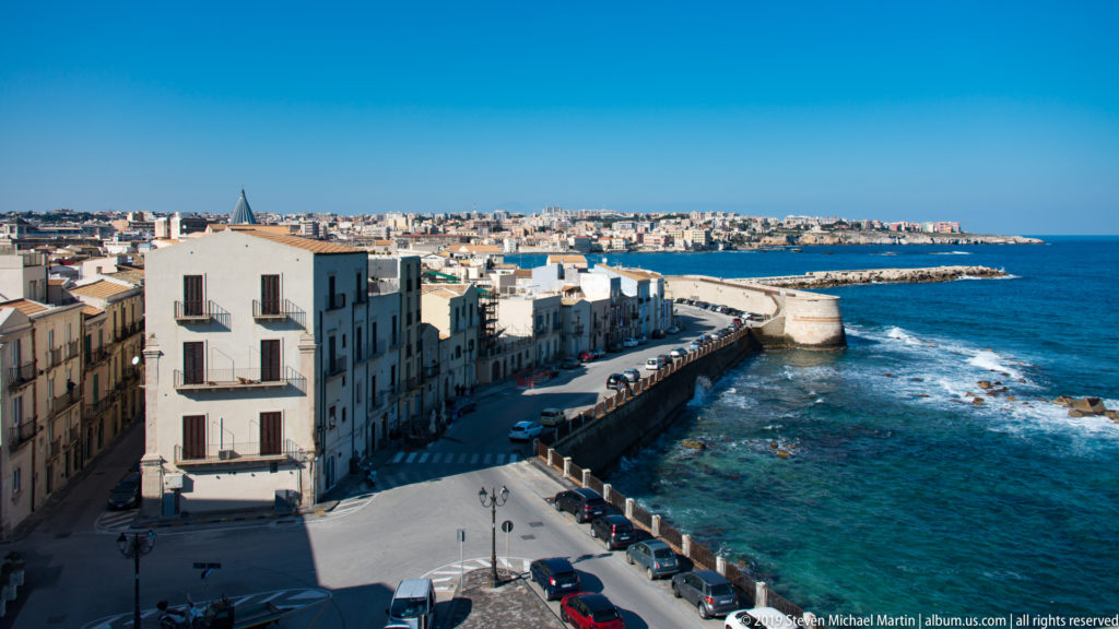 Elevated panorama of Ortigia’s waterfront buildings, coastal road, seawall, rocky shallows, and bright blue sea.