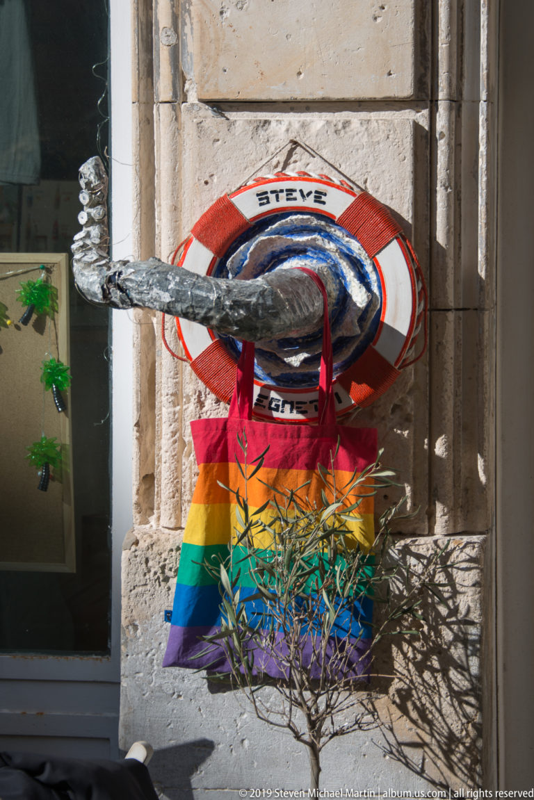 Red-and-white life ring labeled Steve above a rainbow-striped bag and metallic decoration on a pale stone wall.
