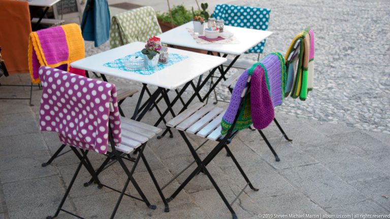 White café tables with colorful polka-dot and crocheted chair covers arranged on a stone-paved pedestrian area.