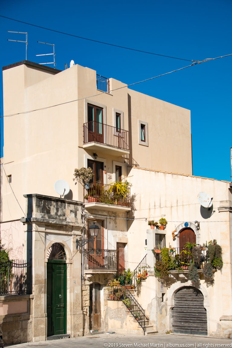 Whitewashed Ortigia residence with balconies, exterior steps, green doors, satellite dishes, and many potted plants.
