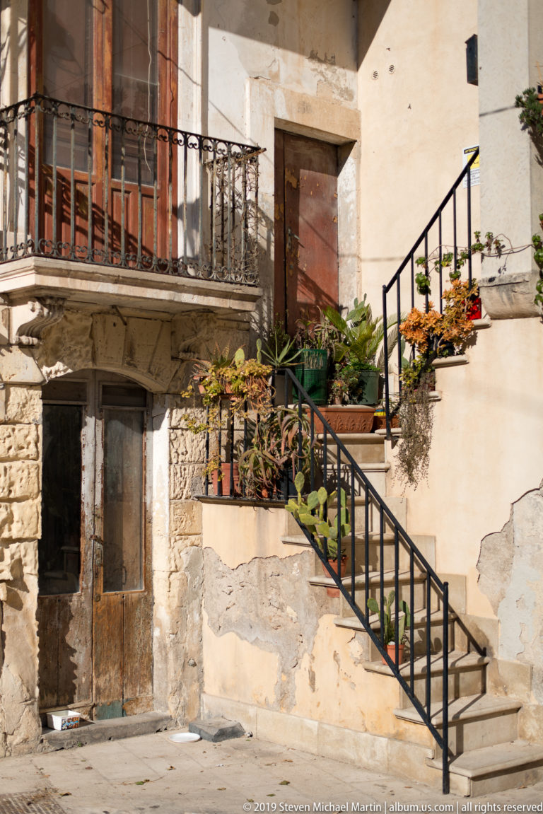 Weathered residence with exterior stair, iron railings, balcony, potted plants, exposed stone, and patched plaster.
