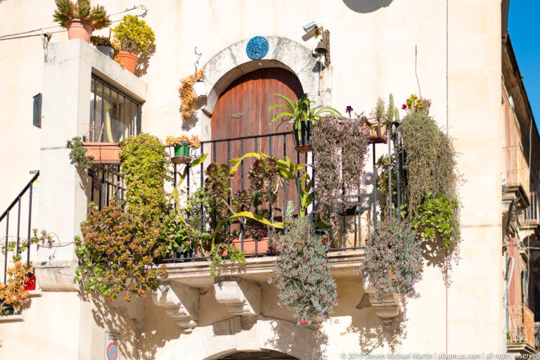 Sunlit residential balcony crowded with potted plants above a stone-framed arched wooden doorway in Ortigia.