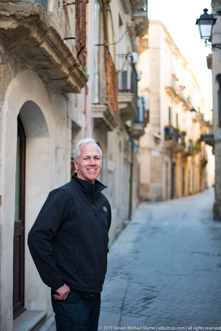 Smiling man in a dark jacket standing on a narrow Ortigia street with historic buildings blurred behind him.