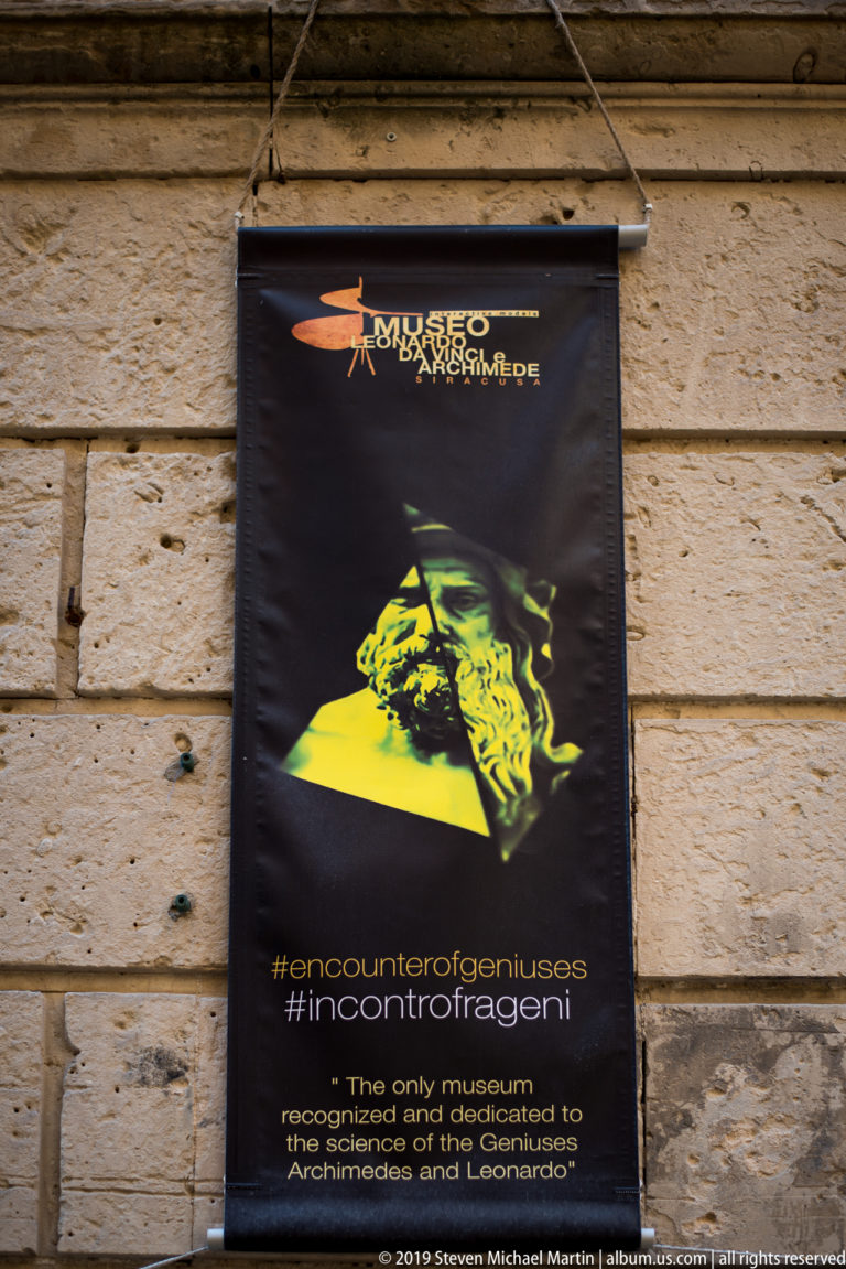 Black museum banner for Leonardo and Archimedes mounted on a pale rusticated stone wall.