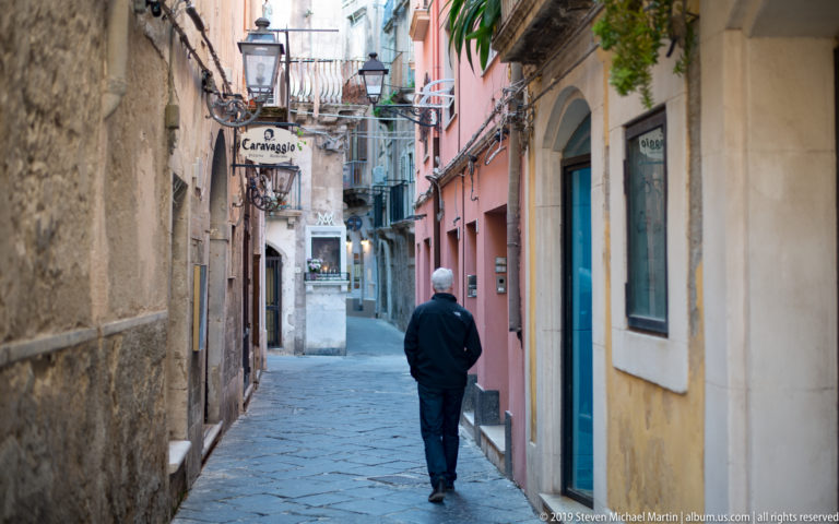 Person walking down a narrow paved Ortigia lane lined with stone and colorful stucco buildings, balconies, and signs.