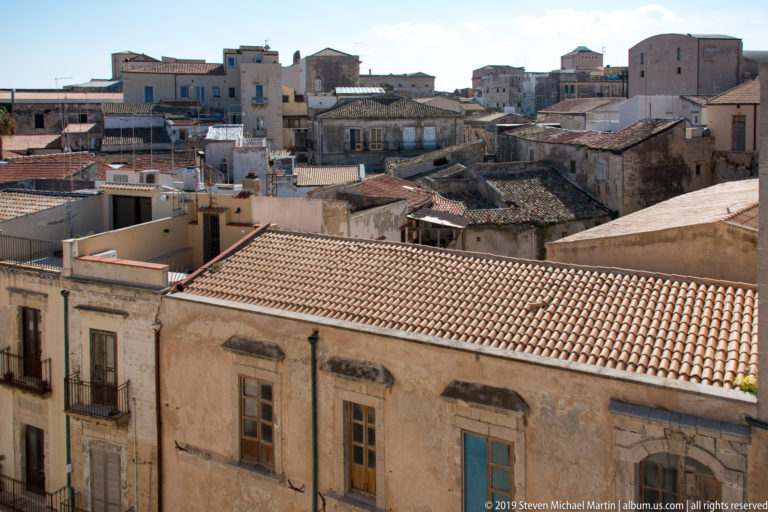 Elevated view across dense Ortigia rooftops with tile roofs, terraces, weathered façades, and closely packed buildings.