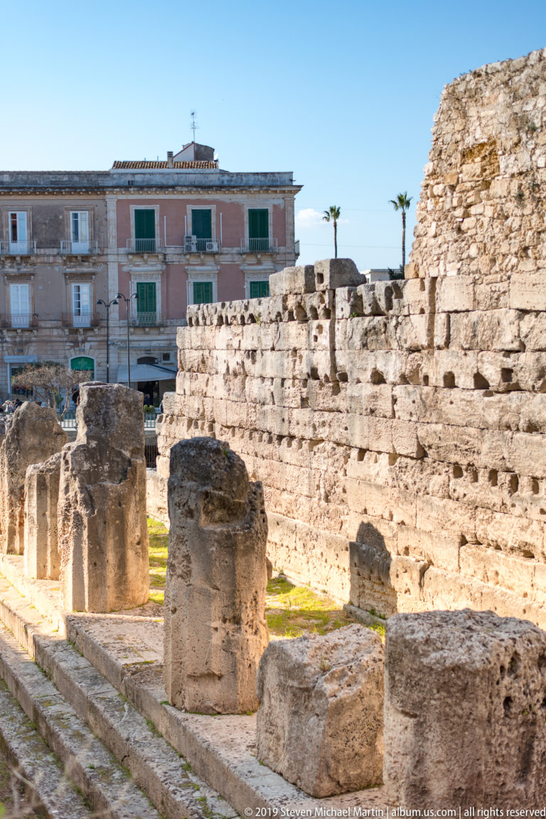 Truncated ancient columns beside layered limestone wall courses at the Temple of Apollo, with residences beyond.