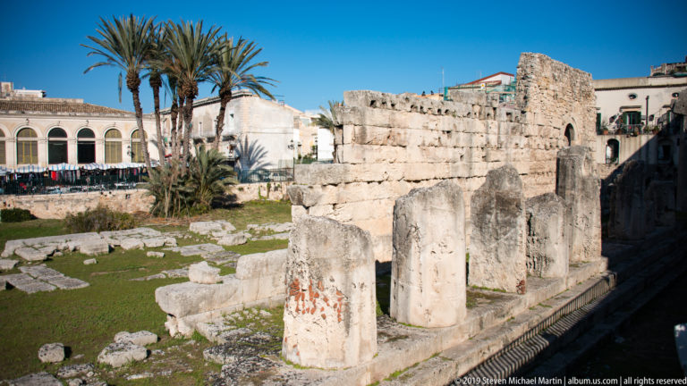 Large weathered column drums and ancient masonry at the Temple of Apollo, with palms and city buildings behind.