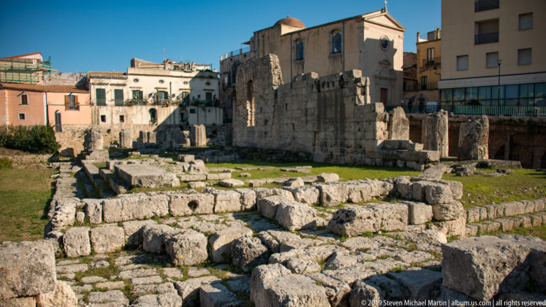 Low limestone foundations and standing Temple of Apollo remains extending toward surrounding buildings in central Ortigia.