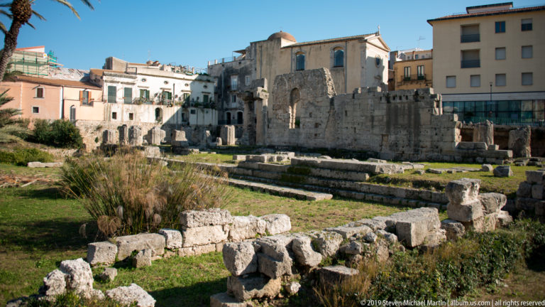 Temple of Apollo ruins with limestone blocks and standing wall sections surrounded by modern buildings in Ortigia.