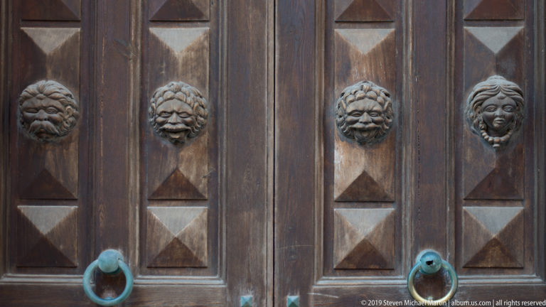 Dark paneled wooden door with four carved lion masks, geometric panels, and two large metal ring handles.