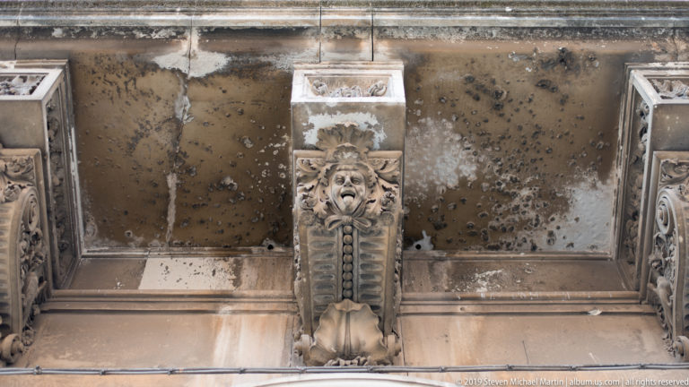 Weathered carved stone corbel with a grotesque face and vertical ornament beneath an aged architectural surface.
