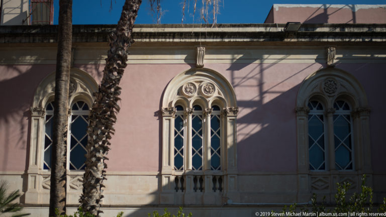 Three ornate pointed windows with pale stone tracery on a pink wall, partly crossed by a palm trunk.