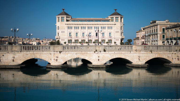 Pale multi-arched Ponte Umbertino crossing calm water with a large white building rising beyond it.