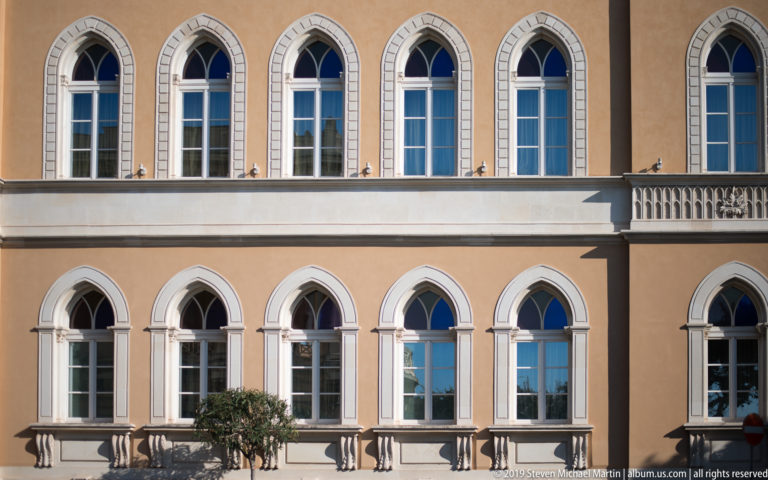 Ocher façade with two rows of pointed-arch windows, pale surrounds, a stone balcony, and a small tree.