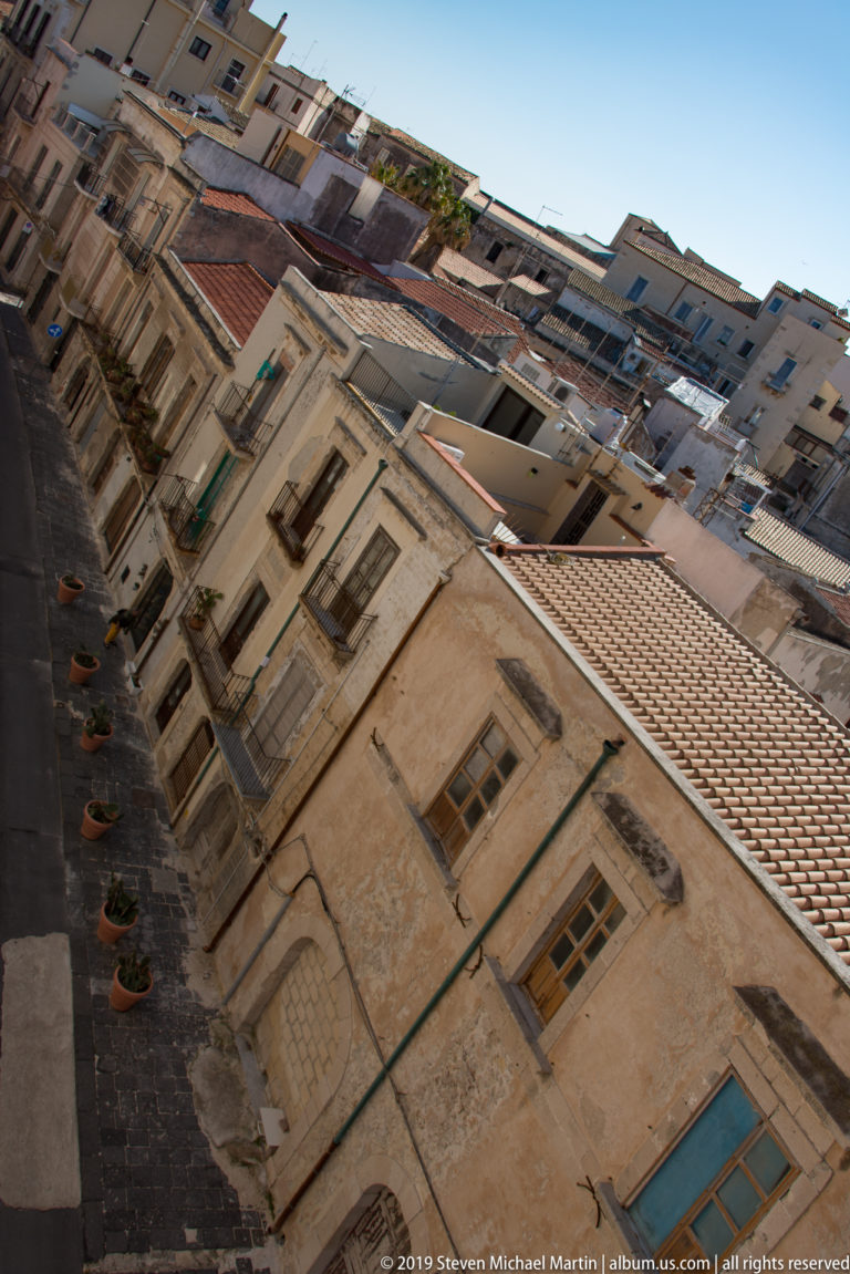 Tilted view down a narrow Ortigia street lined with pale historic façades, shuttered windows, balconies, and tiled roofs.
