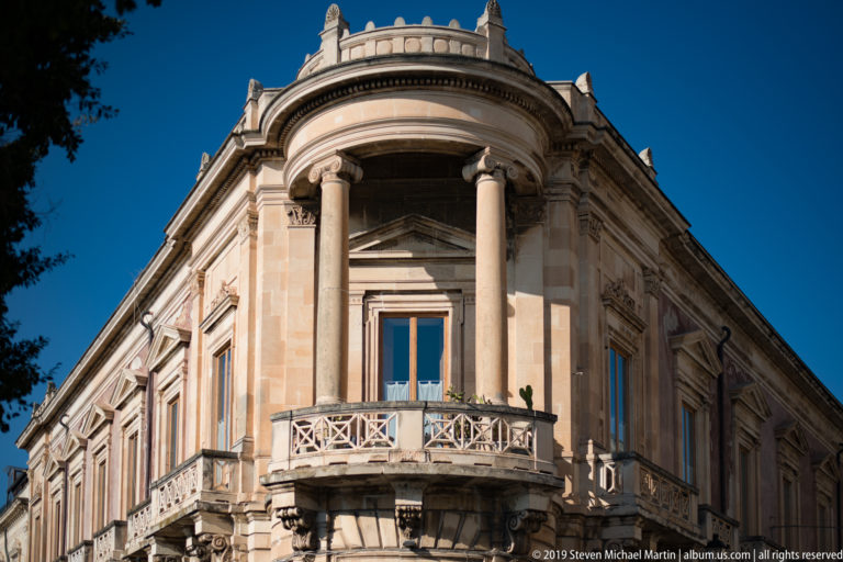 Rounded corner bay with paired columns, curved stone balcony, and tall windows on a pale historic building.