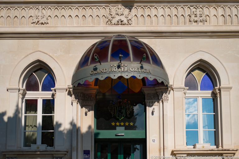 Grand Hotel Ortigia entrance with colorful curved canopy between two pointed-arch windows and decorative stonework.