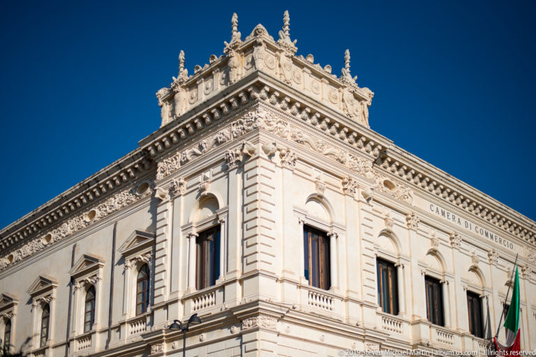 Ornate pale-stone Camera di Commercio corner with arched windows, pilasters, carved cornice, and decorative parapet.