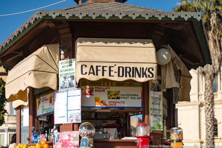 Small caffè and drinks kiosk crowded with menus, snacks, dispensers, and colorful merchandise around its service window.