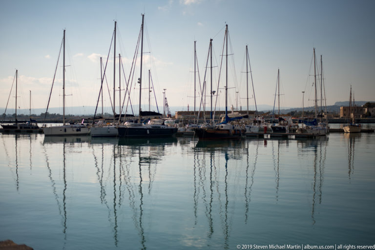 Moored sailboats and tall masts reflected in still marina water beneath a clear sky.