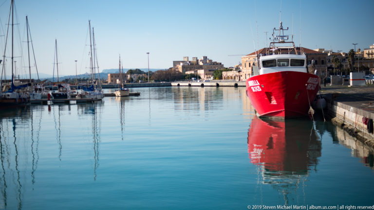 Red workboat moored at a stone quay, reflected in calm blue harbor water with sailboats behind.