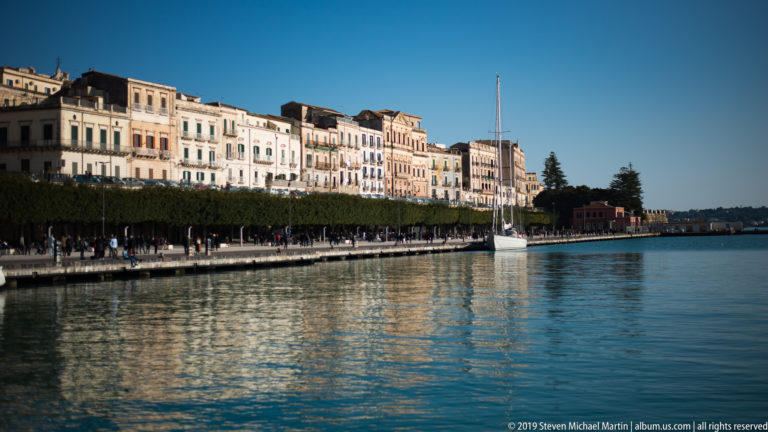 Calm harbor beside Ortigia waterfront buildings, a tree-lined promenade, and a white sailboat moored near shore.