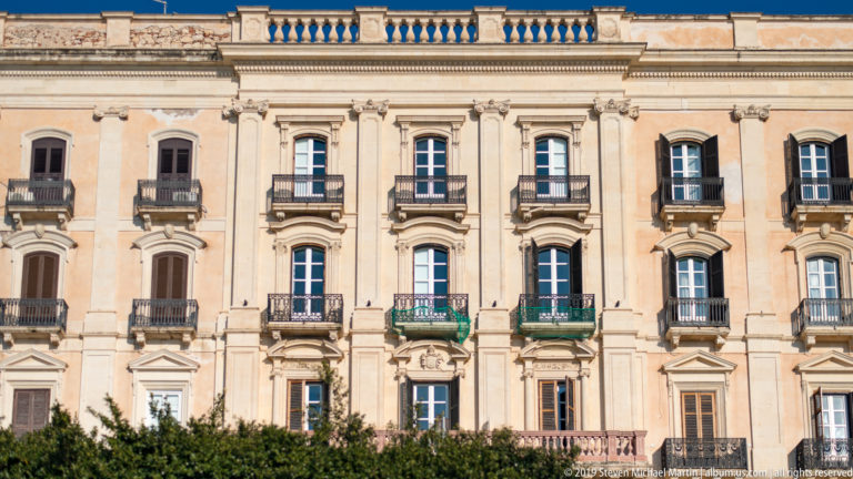 Pale historic façade with rows of windows, iron balconies, pilasters, and a balustraded roofline behind trees.