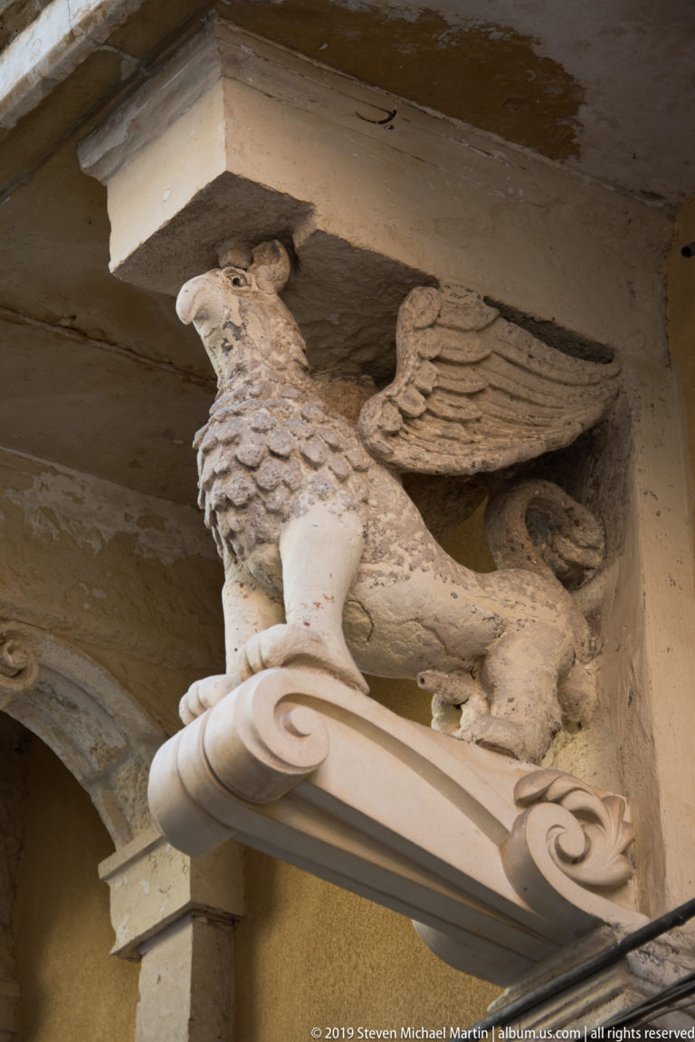 Carved pale-stone winged griffin crouching on a scroll-shaped architectural support beneath a projecting ledge.