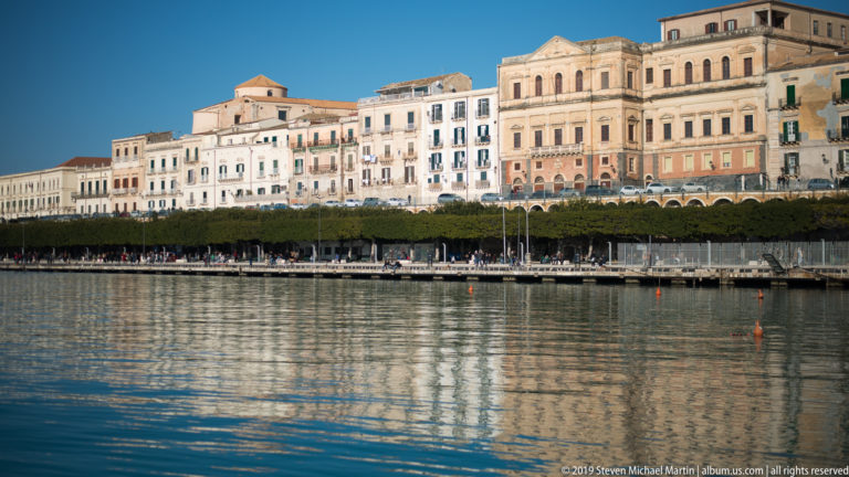 Row of historic multi-story buildings and a tree-lined promenade reflected in calm waterfront water at Ortigia.
