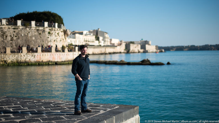 Man standing on a stone waterfront platform with calm sea and Ortigia’s historic seawall behind him.