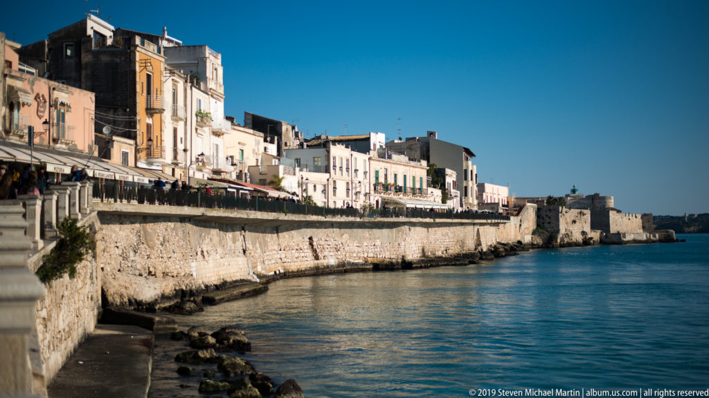 Historic buildings line a high pale stone seawall beside calm blue water on the Ortigia waterfront.