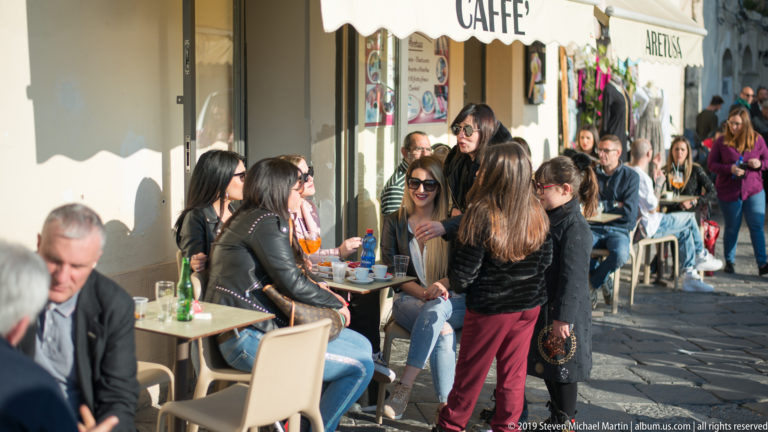 People sit at outdoor café tables along a sunlit Ortigia street while pedestrians pass on the stone pavement.
