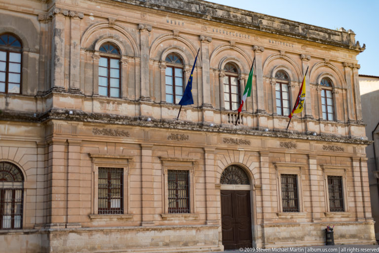 Two-story ornate limestone civic façade with arched windows, central doorway, and European, Italian, and regional flags.