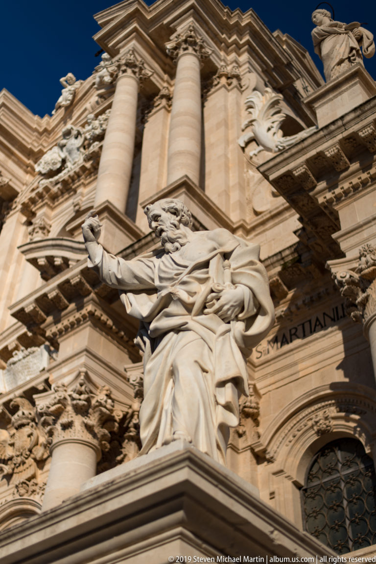 Low-angle detail of a robed stone saint figure among columns and elaborate Baroque façade carvings.