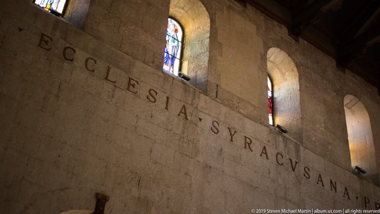 Oblique view of a Latin inscription carved across pale stone masonry beneath tall stained-glass windows.