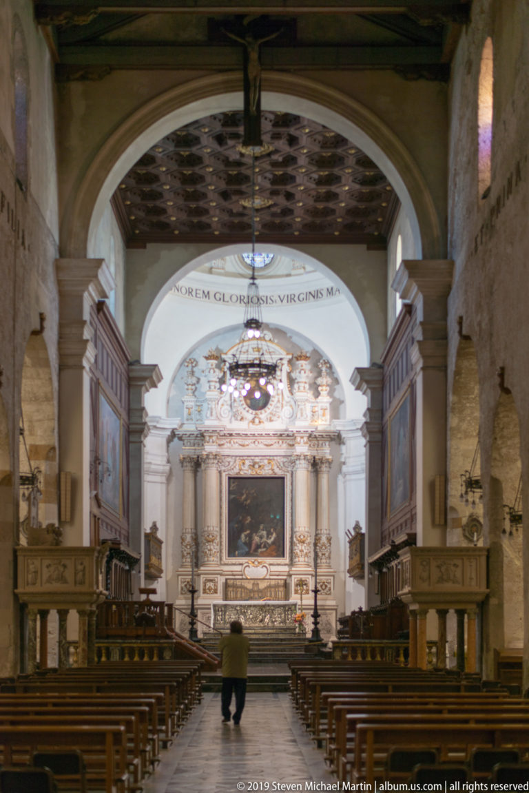 Centered cathedral nave with pews, a single visitor, dark choir enclosures, chandelier, and ornate altar beyond.