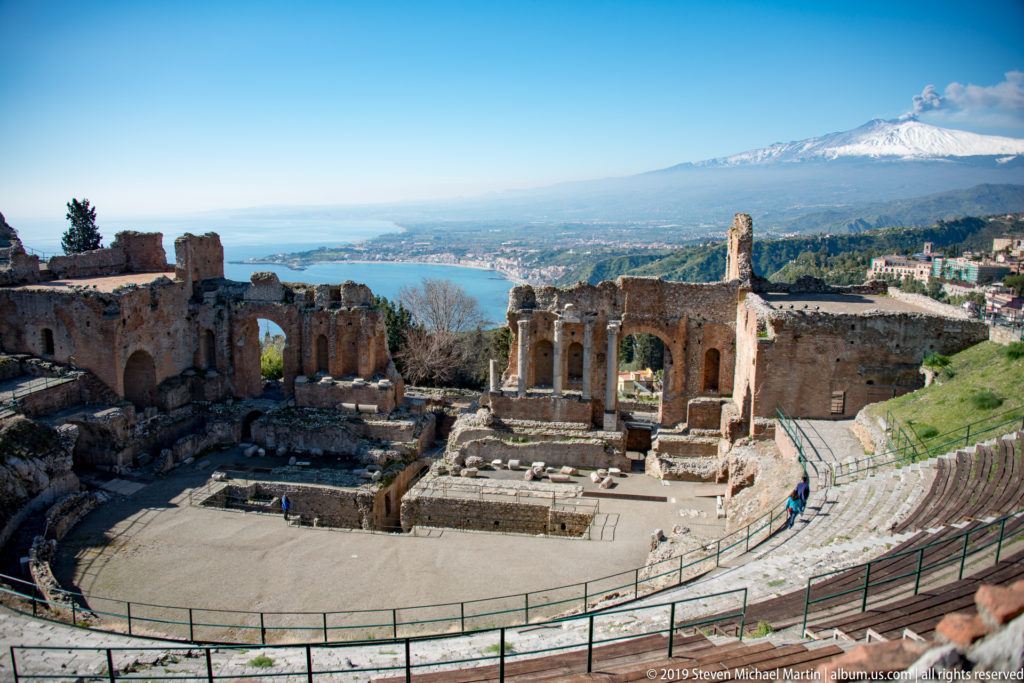 Ancient theatre seating and stage ruins overlooking the coast, with snow-covered Mount Etna rising in the distance.