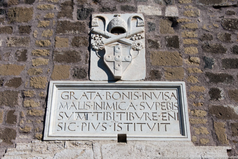 Carved coat of arms above a rectangular Latin inscription mounted on a rough stone fortress wall.