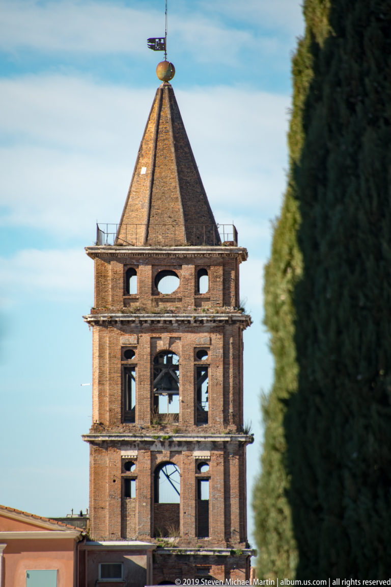 Upper portion of a fortress tower with open brick stage, steep pyramidal roof, and weather vane against blue sky.