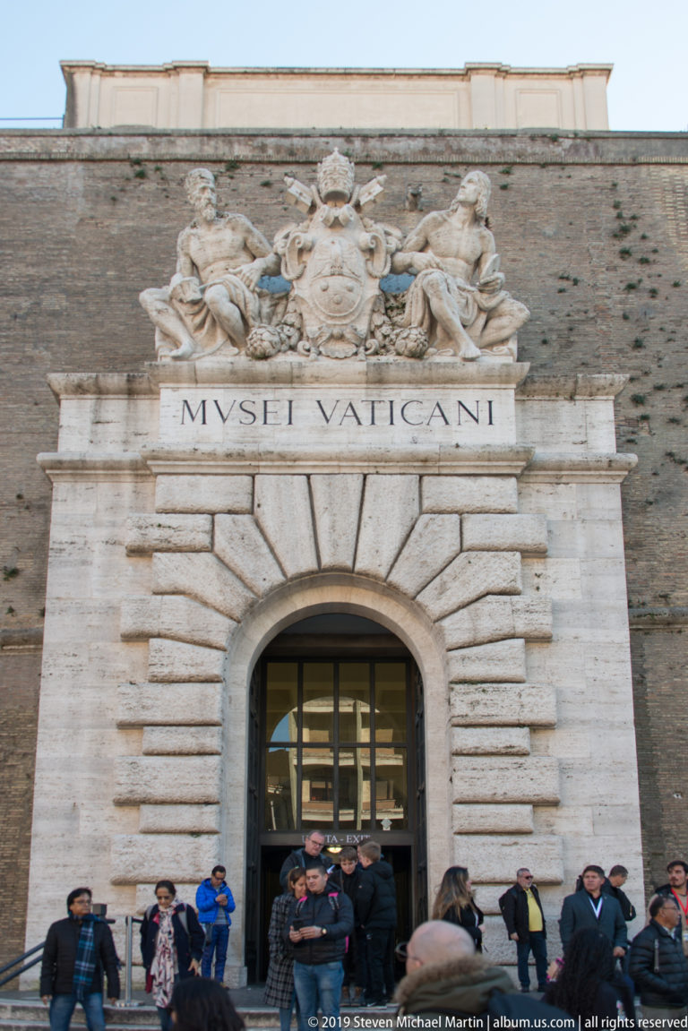 Visitors outside the Vatican Museums entrance beneath the MUSEI VATICANI inscription, papal crest, and tall rusticated arched portal.